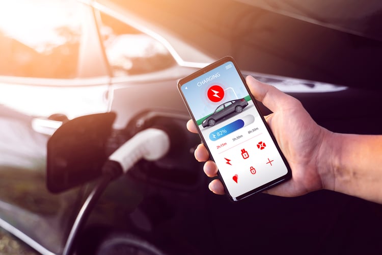 A closeup of a hand holding a smartphone and showing some enabled smart charging features. In the background a car is plugged in and charging.