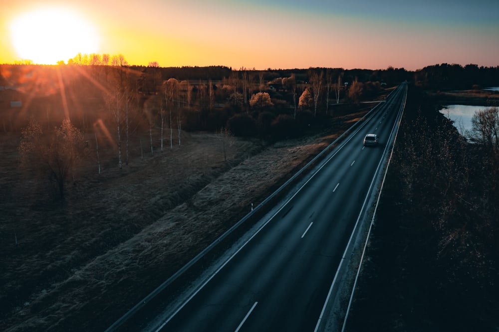 A car is driving on a lonely road in the countryside at sunset.