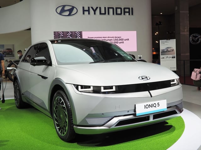 A white Hyundai IONIQ 5 on display at a promotional event with a large Hyndai logo on the wall behind.
