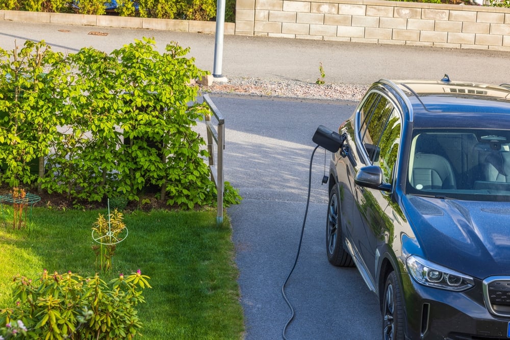 Een zwarte elektrische SUV, van het merk BMW, staat op de oprit van een huis te laden in het zonnetje.