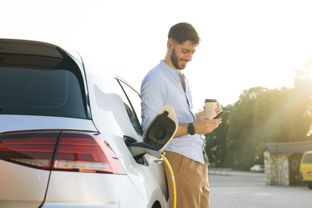 A smiling man is looking at his smartphone while holding a cup of coffee in the other hand on a sunny day. He is leaning on an electric car that is charging.
